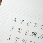 A close-up of a lined paper sheet showing a sample alphabet in elegant cursive script, with letters A through M visible in dark green, brown, and black ink.