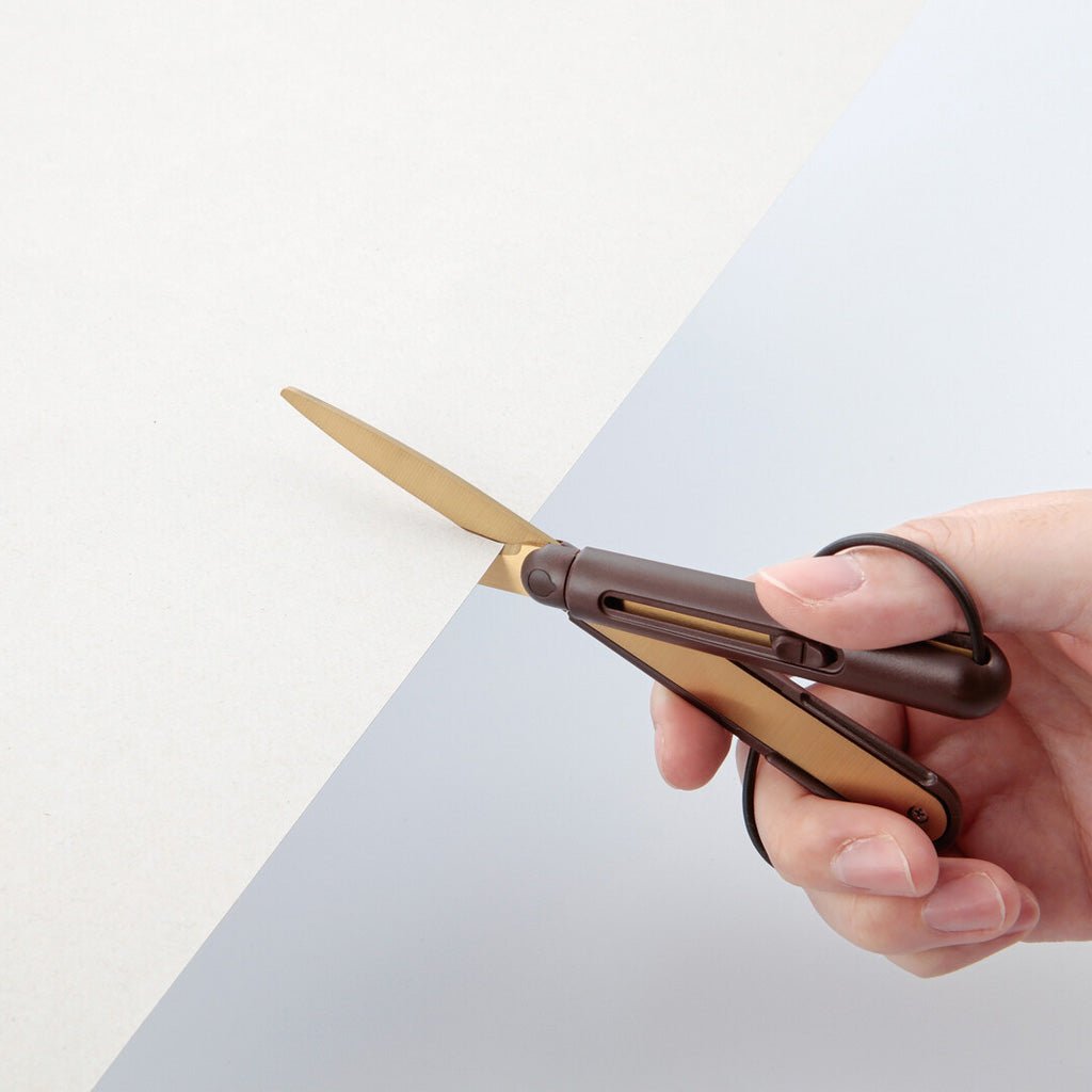 A hand holding a pair of scissors with a dark brown handle and gold-toned blades, shown against a split white and light gray background.