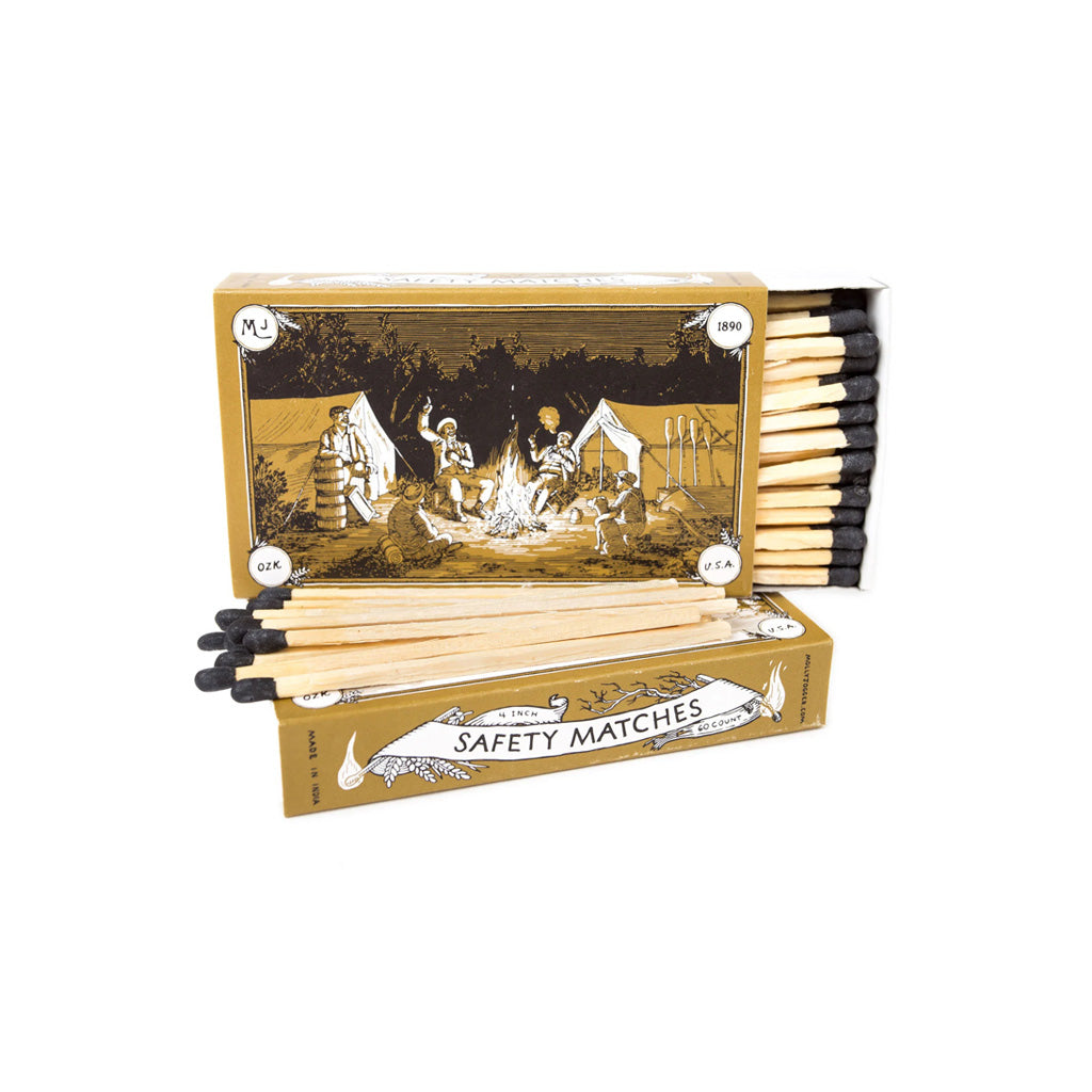 A vintage-style matchbox labeled 'SAFETY MATCHES' with illustrated campfire artwork on the box and several wooden matches with black tips arranged in front, shown on a plain white background.
