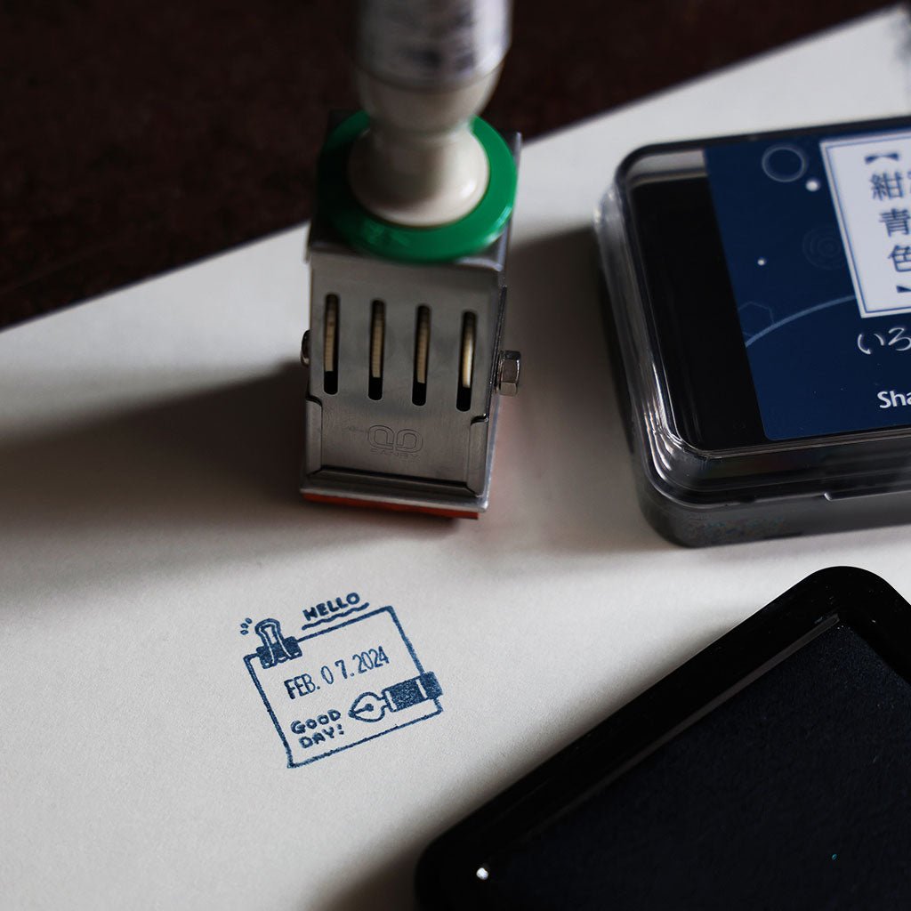 An imprint from a rotary stamp that shows the date, surrounded by a border decorated with a hand drawn binder clip and fountain pen, as well as the words Hello and Good Day!