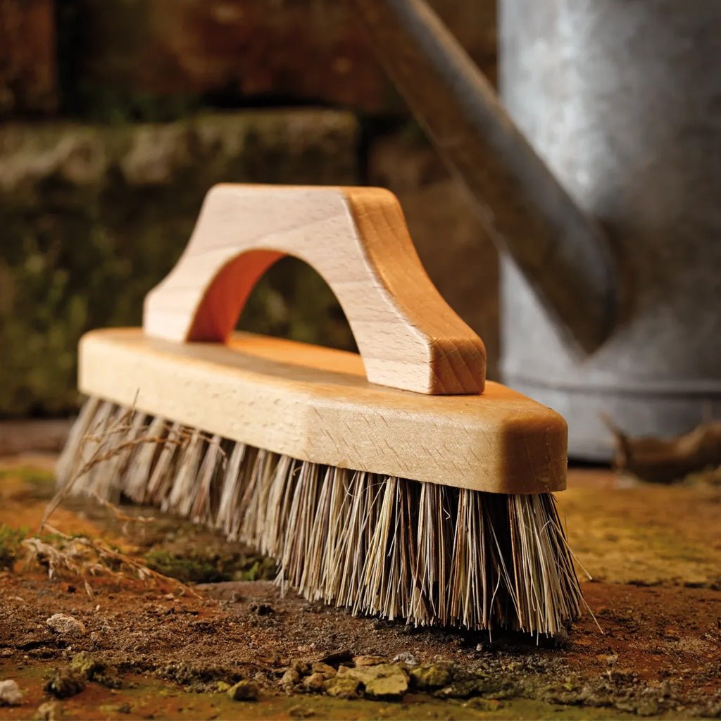 A close-up of a wooden brush with natural bristles, resting on soil with a rustic metal watering can in the blurred background.