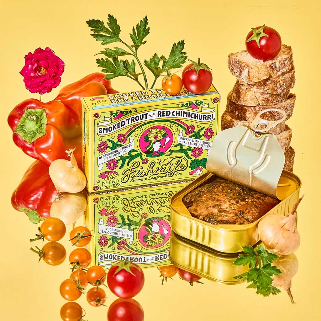 Packaging of tinned Fishwife Smoked Trout with Red Chimichurri on colorful background next to produce and bread