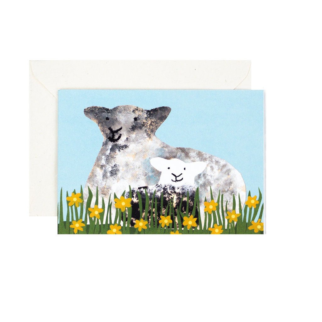 A greeting card featuring an illustration of a sheep with a lamb, surrounded by yellow flowers, with a blue sky in the background. The card is set against a white envelope.