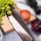 Knife with '3 Claveles' branding holding green vegetable against a blurred kitchen background.