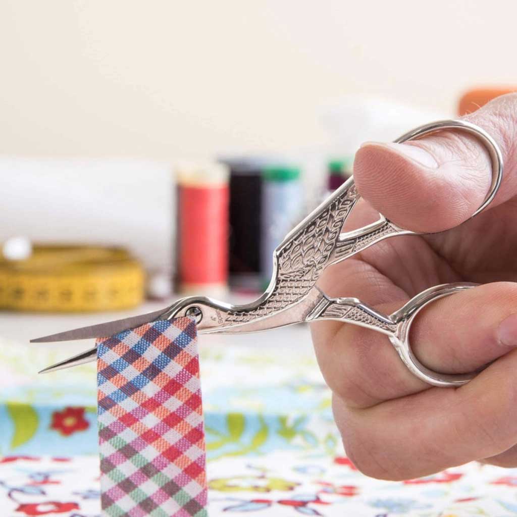 Hand holding a pair of silver scissors cutting colorful fabric with sewing materials in the background.