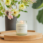 Candle labeled 'Sunday Morning' on a wooden tray with flowers in the background