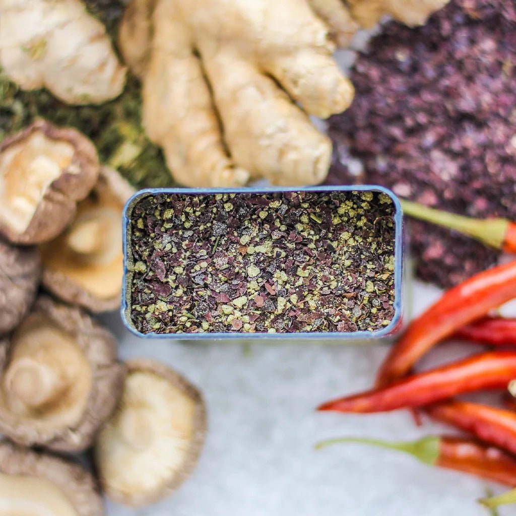 Dried herbs in a small container surrounded by mushrooms, ginger, and red chili peppers on a marble surface.