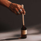 Hand holding a bottle of fragrance with wooden diffuser sticks against a dark background