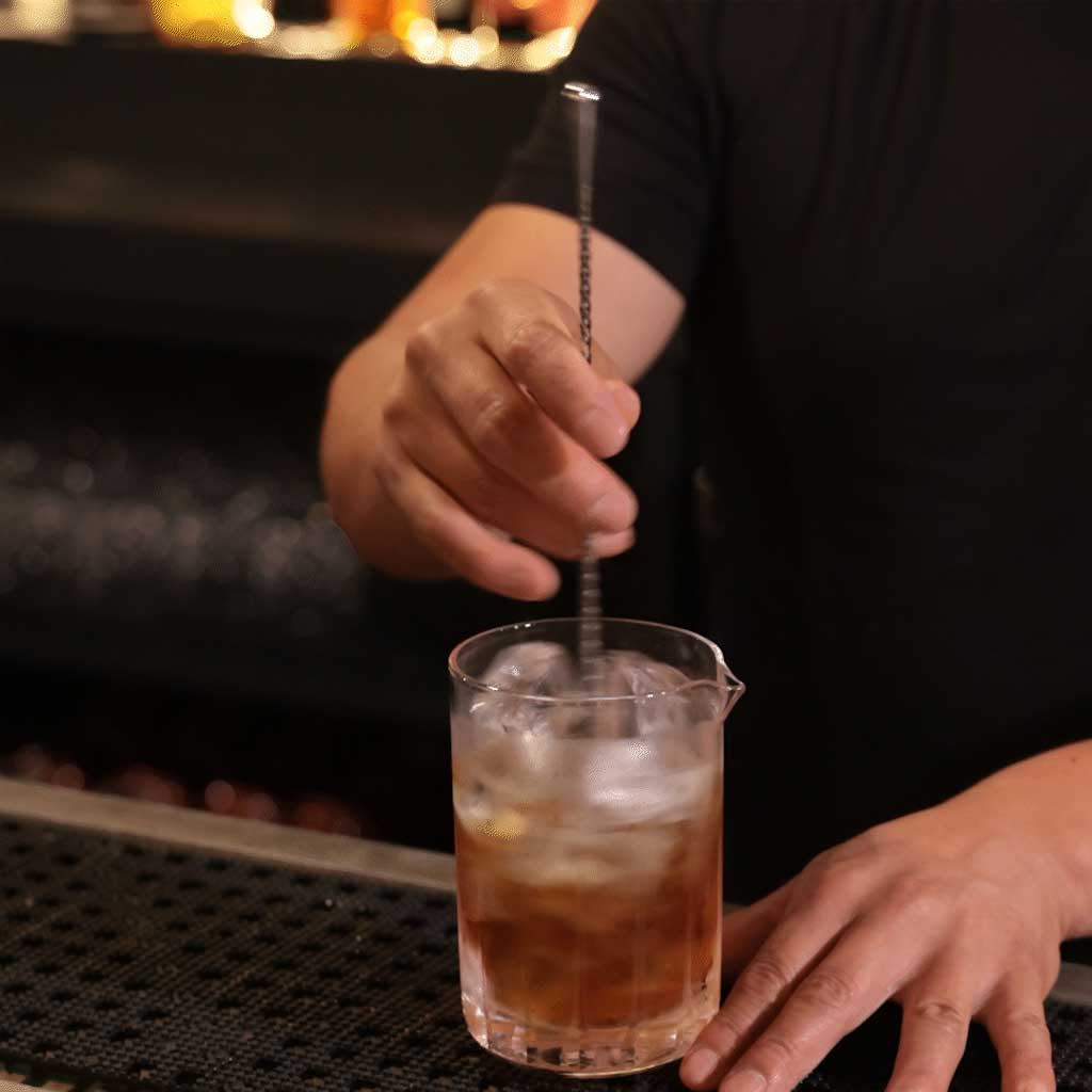 Person stirring a cocktail with ice in a glass at a bar.