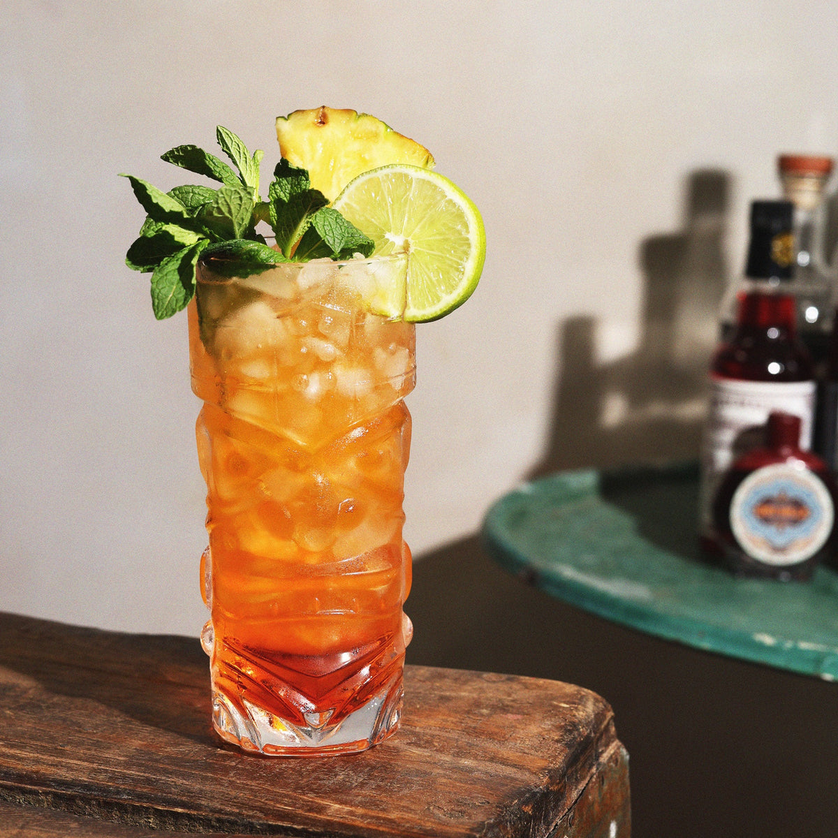 A tall glass with a tiki design is filled with an amber colored drink garnished with mint and tropical fruits. 