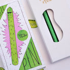 A green metallic pen in a white window box packaging, shown next to its illustrated outer box featuring a pickle design and the brand name 'Tom's Studio'.