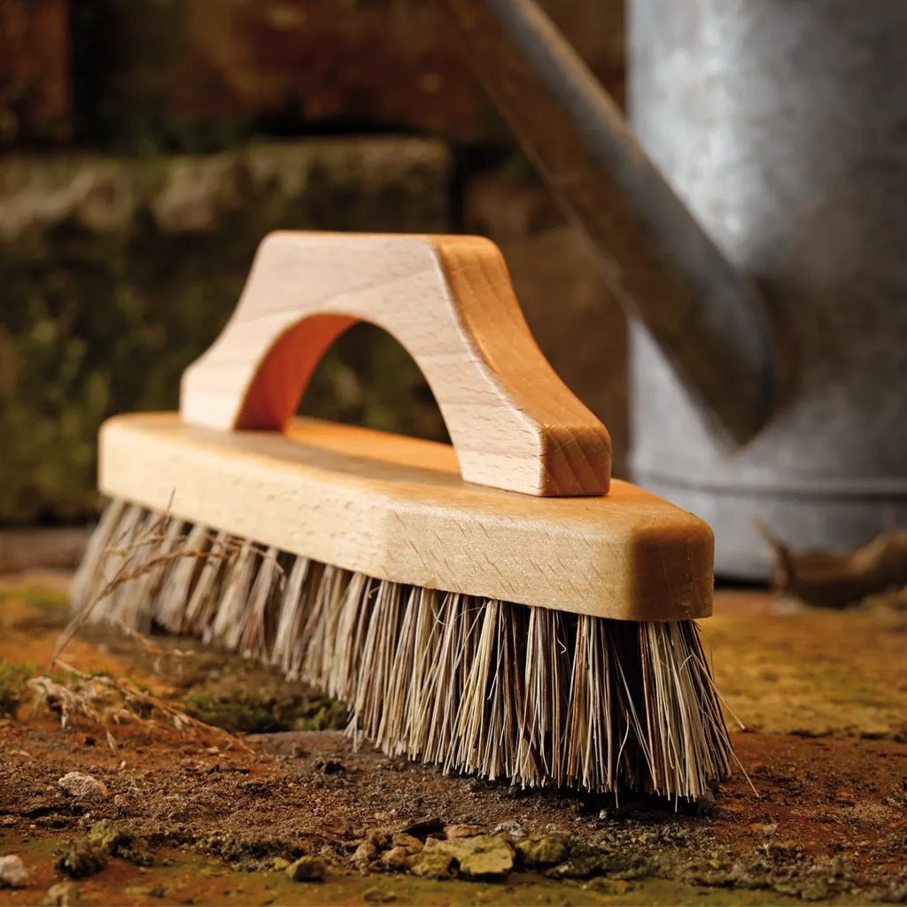 A close-up of a wooden brush with natural bristles, resting on soil with a rustic metal watering can in the blurred background.