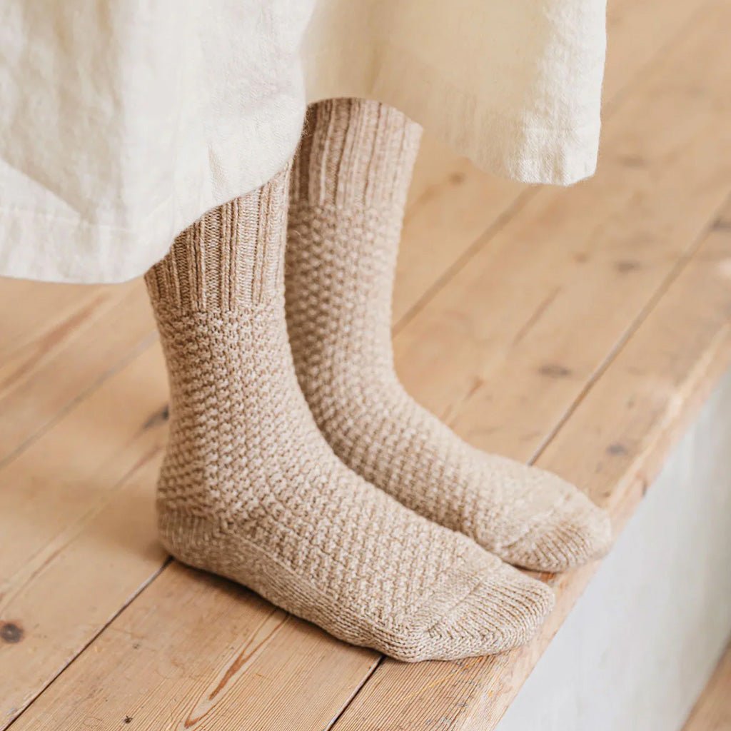 A pair of beige chunky knit socks worn on feet, shown with the hem of a white dress at the top, set against a light wooden plank floor.