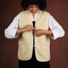 A model wearing a natural woolen vest over a white shirt and black jumper. She is fastening the toggle closure.