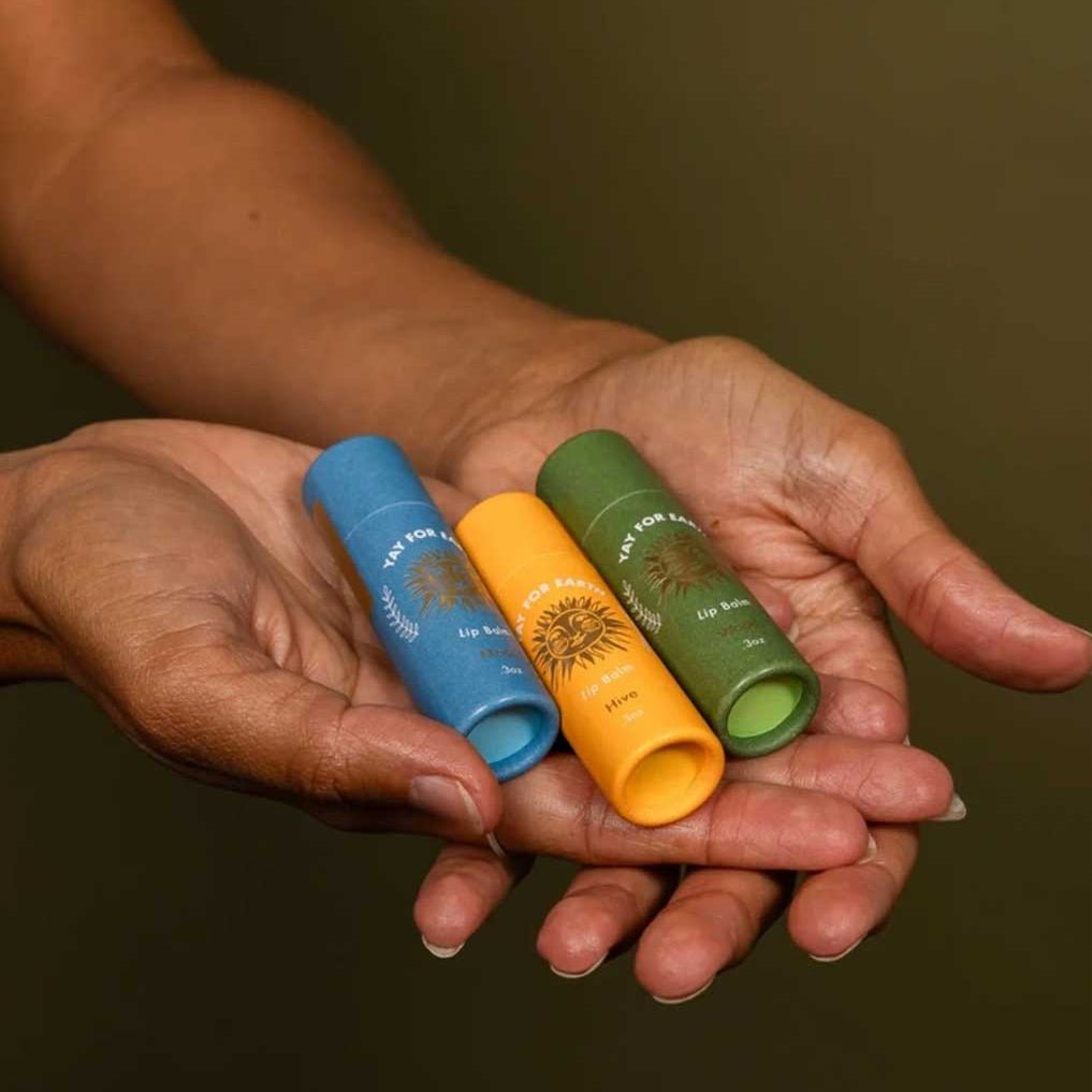Three lip balm tubes in blue, orange, and green held in a person's hands against a dark background.