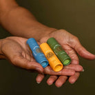 Three lip balm tubes in blue, orange, and green held in a person's hands against a dark background.