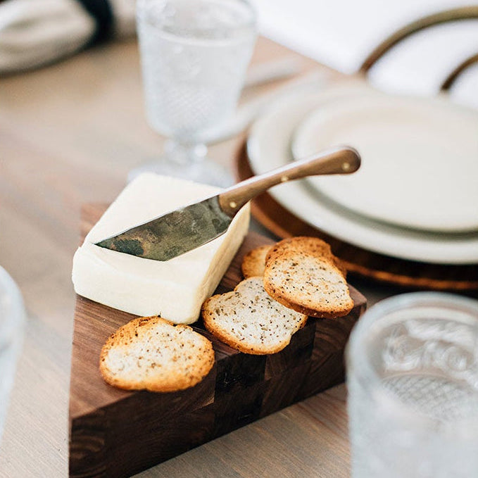 Hand-forged stainless steel spreader knife with walnut handle in block of cheese on charcuterie board on table