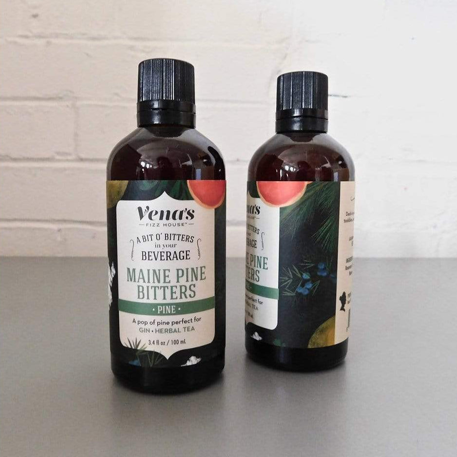 Two bottles of Maine Pine Bitters by Vena's on a light surface with a white brick wall background.